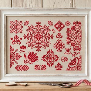 Può includere: Un ricamo a punto croce incorniciato che presenta una collezione di intricati motivi geometrici e floreali rossi su uno sfondo in tessuto beige chiaro. L'opera è racchiusa in una cornice di legno bianco con finitura anticata.