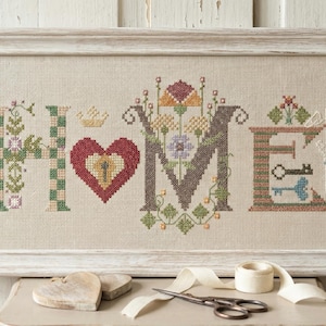 May include: Cross-stitch artwork spelling "HOME" in a white framed piece. The letters are decorated with floral and geometric patterns. The "H" is green and white checkered, the "O" is a red heart, and the "E" has keys. A wooden heart, scissors, and tape are on the table.