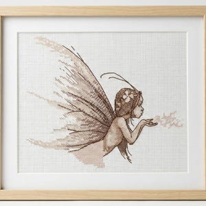 May include: Framed cross-stitch art featuring a fairy with large, detailed wings and a floral crown. The fairy is blowing a puff of what appears to be glitter or dust. The artwork is in shades of brown and beige, set in a light wood frame.