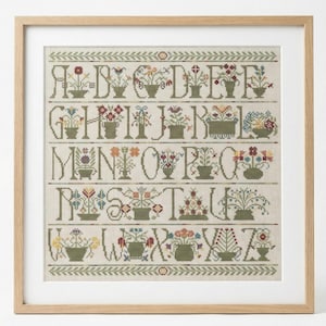 Puede incluir: Muestra de punto de cruz enmarcada con el alfabeto, cada letra representada por una flor en maceta. El bordado utiliza tonos verdes, rojos, amarillos y azules sobre un fondo neutro. El marco de madera añade un toque clásico y elegante.