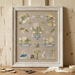 Op de afbeelding: Een ingelijste kruissteek sampler met een crèmekleurige achtergrond. Het ontwerp bevat bloemstukken, vogels en een klein huis. Het witte frame staat op een houten oppervlak met een schaar, lint en houten harten.