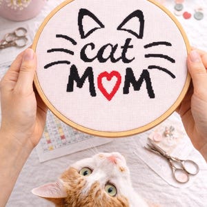 May include: A cross-stitch embroidery hoop with the words "cat MOM" in black and red thread. The design includes cat ears and whiskers. A calico cat looks up at the hoop. The hoop is held by two hands.