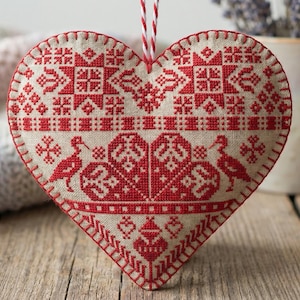 May include: Heart-shaped, embroidered ornament with a red and white folk art design. The ornament features geometric patterns and bird motifs. It hangs by a red and white string. The background includes a soft blanket and a mug with lavender.