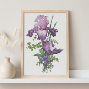 Pu&ograve; includere: Una stampa ad acquerello incorniciata di fiori di iris viola con fogliame verde e piccole bacche blu. L'opera d'arte &egrave; in una cornice di legno chiaro ed &egrave; esposta su una mensola bianca accanto a un vaso color crema con fiori secchi.