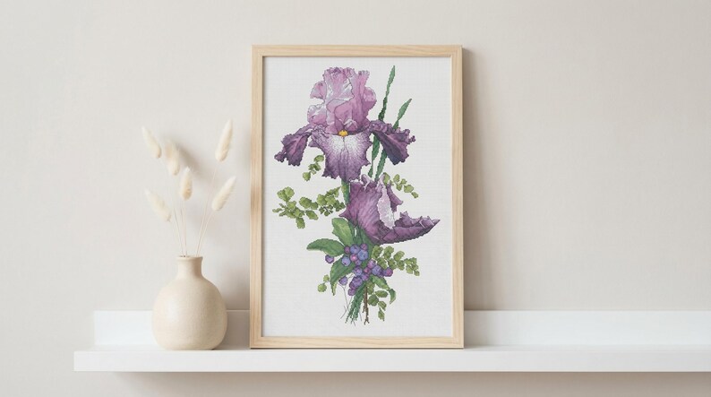 Pu&ograve; includere: Una stampa ad acquerello incorniciata di fiori di iris viola con fogliame verde e piccole bacche blu. L'opera d'arte &egrave; in una cornice di legno chiaro ed &egrave; esposta su una mensola bianca accanto a un vaso color crema con fiori secchi.