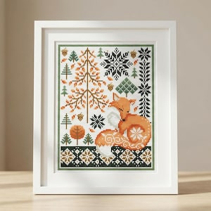 以下が含まれることがあります： 額縁に入ったクロスステッチのアートワーク。オレンジと白のキツネのデザインで、木、雪の結晶、緑、茶色、白の模様に囲まれています。白いフレームに飾られています。