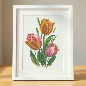 Op de afbeelding: Een ingelijst kruissteekkunstwerk met drie tulpen in tinten oranje, roze en geel, met groene bladeren. Het kunstwerk is in een witte lijst, op een houten oppervlak.