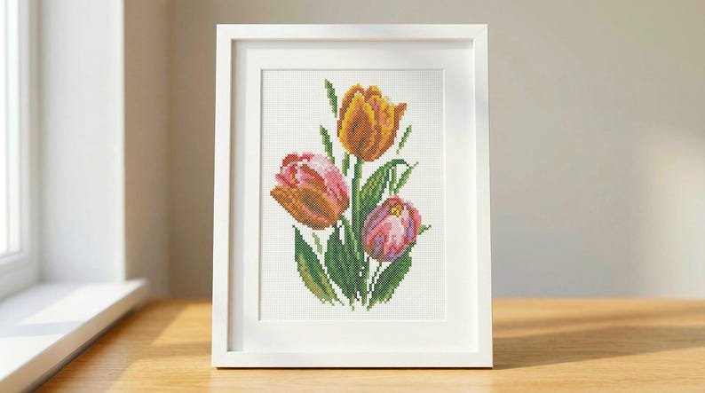 Op de afbeelding: Een ingelijst kruissteekkunstwerk met drie tulpen in tinten oranje, roze en geel, met groene bladeren. Het kunstwerk is in een witte lijst, op een houten oppervlak.