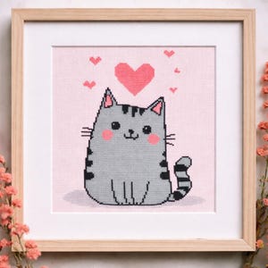 Puede incluir: Un punto de cruz enmarcado con un gato gris con rayas negras, mejillas rosas y un corazón encima de su cabeza. El gato está sobre un fondo rosa con pequeños corazones rosas. El marco es de madera clara.