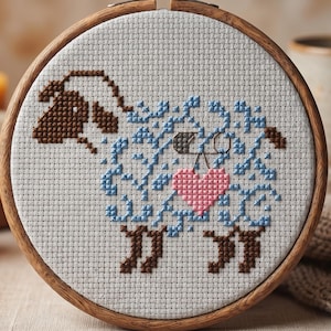 Könnte beinhalten: Ein Kreuzstich-Stickrahmen mit einem Schaf-Design. Das Schaf hat einen braunen Kopf, einen blauen Körper und ein rosa Herz. Die Stickerei befindet sich in einem Holzrahmen. Kerzen und eine Tasse sind im Hintergrund.