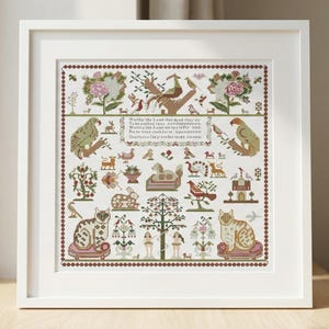 May include: Framed cross-stitch sampler with a white frame. The sampler features a detailed design with various animals, birds, and floral elements in shades of green, brown, and pink. The text is visible in the center. The artwork is displayed on a wooden surface.