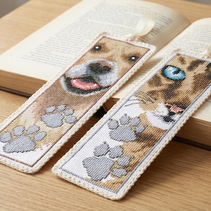May include: Two embroidered bookmarks featuring a dog and a cat. The dog bookmark shows a smiling dog with paw prints, and the cat bookmark has a blue-eyed cat with paw prints. Both have a cream-colored border and a ribbon.