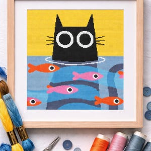 Puede incluir: Imagen de punto de cruz de un gato negro en el agua, rodeado de peces naranjas y rosas. La cabeza y los ojos del gato son visibles. La obra está enmarcada en un marco de madera clara. Suministros de costura en primer plano.