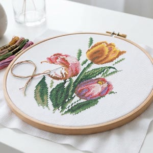 Op de afbeelding: Een borduurring met kruissteek met een bloemenontwerp van tulpen in roze, oranje en geel. De borduursels zijn op witte stof, geplaatst in een houten ring. Borduurgaren in verschillende kleuren en een naald zijn ook aanwezig.