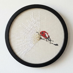 May include: A framed embroidery featuring a red ladybug perched on a branch, set against a white web design. The artwork is enclosed in a round black frame, creating a striking contrast. The background is a neutral off-white.
