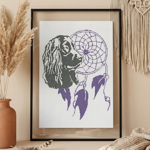 May include: Cross-stitch art featuring a black and gray dog with a purple dreamcatcher. The artwork is framed in a black frame and displayed on a neutral-toned wall. Decorative feathers and a knitted blanket are in the foreground.