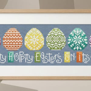 May include: A framed cross-stitch artwork showing six patterned Easter eggs in a row. The text "Hippy Hoppy Easter's ON Its Way" is stitched below the eggs. The background is blue, and the frame is made of wood.
