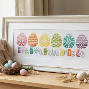 May include: A framed cross-stitch featuring six colorful Easter eggs with intricate patterns and the text "Hiply Hopity Easter's ON Its Day." A plush bunny, a nest with eggs, and decorative eggs are on a wooden table.