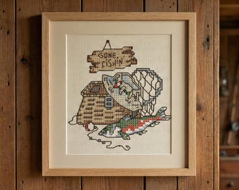 May include: Framed cross-stitch artwork with a fishing theme. The design includes a fish, a tackle box, and the words "Gone Fishin'" in a rustic style. The artwork is framed in a light wood frame.