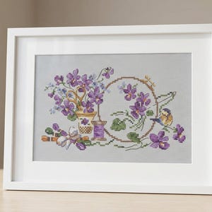 Può includere: Opera d'arte a punto croce incorniciata con un design floreale con fiori viola, un uccellino e rocchetti di filo. L'opera d'arte è in una cornice bianca e poggia su una superficie di legno. La tavolozza dei colori comprende tonalità di viola, verde e marrone.