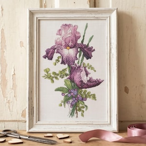 Pu&ograve; includere: Un ricamo incorniciato di un fiore di iris viola con foglie verdi e piccole bacche viola. L'opera d'arte &egrave; in una cornice bianca invecchiata. Un paio di forbici, cuori di legno e un nastro rosa sono sulla superficie di legno.