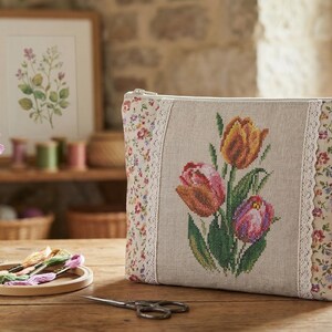 Op de afbeelding: Een beige etui met ritssluiting en een kruissteek tulpenontwerp. De etui heeft bloemmotief zijkanten en een kanten rand. Een vaas met paarse en roze bloemen, borduurgaren en een ingelijste botanische print staan ook op de houten tafel.