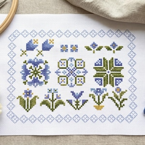 May include: A cross-stitch pattern featuring blue and green floral designs on white fabric. The embroidery is surrounded by a blue geometric border. Accessories include embroidery floss, scissors, and a wooden hoop.