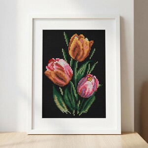 Op de afbeelding: Een ingelijste kruissteek van drie tulpen in tinten oranje, roze en wit tegen een zwarte achtergrond. De bloemen hebben groene stelen en bladeren. Het kunstwerk wordt gepresenteerd in een witte lijst.
