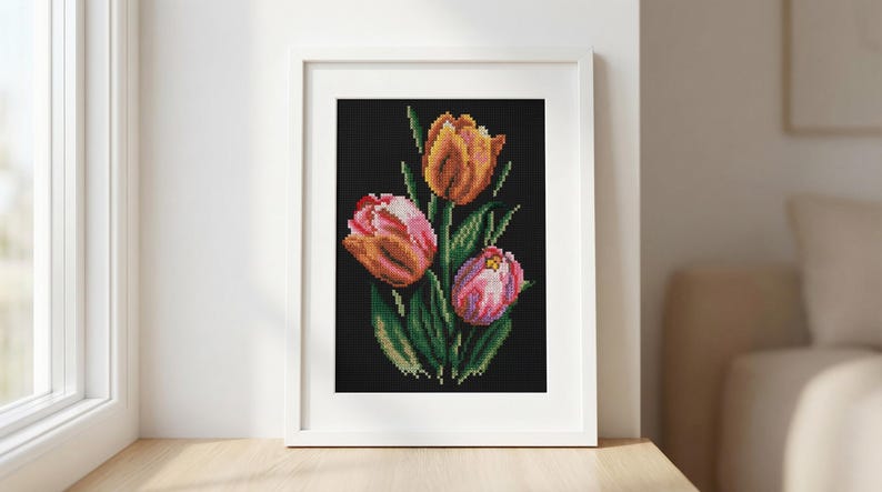 Op de afbeelding: Een ingelijste kruissteek van drie tulpen in tinten oranje, roze en wit tegen een zwarte achtergrond. De bloemen hebben groene stelen en bladeren. Het kunstwerk wordt gepresenteerd in een witte lijst.