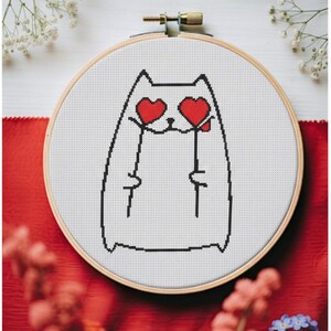 May include: A cross-stitch embroidery in a wooden hoop features a cat with heart-shaped eyes and holding two red hearts. The design is on white fabric, with a black outline. The background includes red fabric and white and blue flowers.