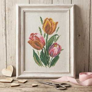 Op de afbeelding: Een ingelijste kruissteek van drie tulpen in tinten oranje, roze en paars, met groene bladeren. Het ingelijste kunstwerk wordt gepresenteerd op een houten oppervlak met houten harten, een schaar en een roze lint.