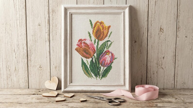 Op de afbeelding: Een ingelijste kruissteek van drie tulpen in tinten oranje, roze en paars, met groene bladeren. Het ingelijste kunstwerk wordt gepresenteerd op een houten oppervlak met houten harten, een schaar en een roze lint.
