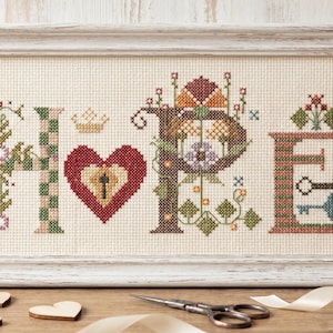 May include: Cross-stitch artwork in a white frame spelling "HOPE". The letters are decorated with floral and geometric patterns, a heart with a keyhole, and a small crown. A pair of scissors and wooden hearts are on the table.