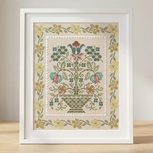 Op de afbeelding: Een ingelijst kruissteek bloemenontwerp. Het kunstwerk toont een centrale vaas die overloopt van bloemen in tinten groen, blauw, rood en perzik. Het ontwerp is omlijst met een witte rand en een bloemenpatroon in geel en groen.