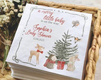 May include: A pile of white paper napkins with a baby shower design. The napkins show a forest scene with a deer, hedgehog, and rabbit. Text reads "a merry little baby is on the way" and "Sophia's Baby Shower 12.08.2025".