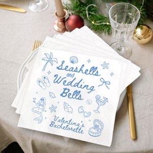 May include: White paper napkins with blue illustrations and text. The napkins feature drawings of seashells, a mermaid, and other beach-themed images. The text reads "Seashells and Wedding Bells, Valentina's Bachelorette."