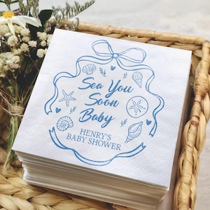 May include: A stack of white paper napkins with a blue nautical design. The design includes seashells, a starfish, and the text "Sea You Soon Baby" and "Henry's Baby Shower". The napkins are in a woven basket.