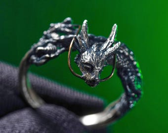 May include: A silver-toned ring featuring a detailed dragon design. The dragon's head is prominent, with intricate scales and horns. The ring has a dark, oxidized finish, and a gold-colored accent encircles the dragon's snout. The ring is set against a green background.