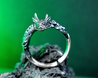 May include: A silver dragon ring with intricate detailing. The dragon's head is at the top, with its body forming the band. The ring is set against a dark, textured surface with a green background.