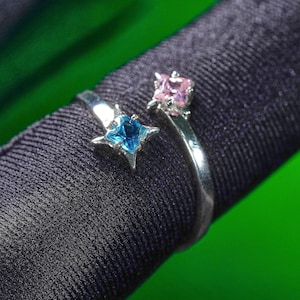 May include: A silver-toned ring with an open design, featuring a blue star-shaped gemstone and a pink square gemstone. The ring is set against a dark fabric background with a green backdrop.