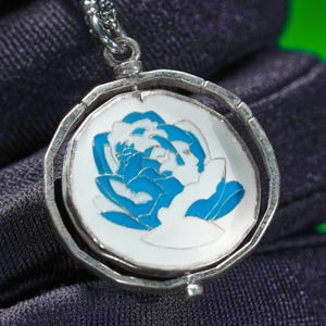 Op de afbeelding: Een zilverkleurige hangerketting met een cirkelvormige bedel met een blauw-wit bloemmotief. De hanger heeft een draaimechanisme en hangt aan een fijne ketting. De achtergrond is een donkerblauwe stof.