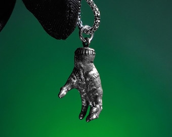 May include: A silver-colored pendant in the shape of a hand, suspended from a chain. The hand pendant is detailed with visible stitching and is set against a gradient green background.