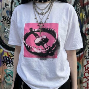 May include: A white short-sleeve t-shirt featuring a graphic print of a black bear trap centered on a pink square background. The shirt is paired with a layered silver-tone chain necklace set that includes multiple cross pendants and a spiked choker.