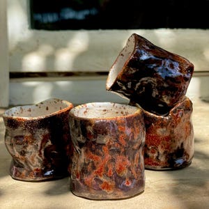 Puede incluir: Cuatro tazas de cerámica hechas a mano con un diseño orgánico texturizado. Las tazas son de color marrón oscuro con detalles en rojo anaranjado y blanco. El interior de cada taza es blanco. Las tazas están apiladas.