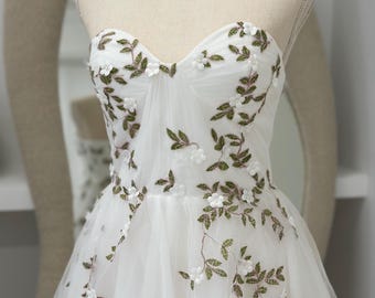 May include: A strapless white wedding dress with floral embroidery. The bodice features delicate white flowers and green leaves, with a tulle skirt. The dress is displayed on a mannequin, showcasing its elegant design.