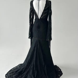May include: A black lace gown with long sleeves and a dramatic train. The dress features a deep V-neckline and a low back. The intricate lace pattern covers the entire dress, creating a flowing silhouette.