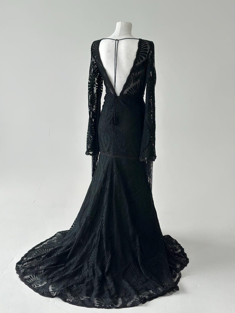 May include: A black lace gown with long sleeves and a dramatic train. The dress features a deep V-neckline and a low back. The intricate lace pattern covers the entire dress, creating a flowing silhouette.
