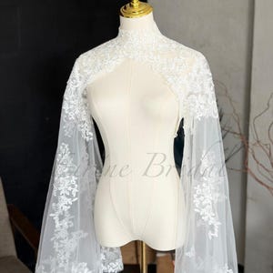 May include: A white bridal bolero featuring a high neckline and long, flared sleeves made of sheer tulle fabric. The garment is adorned with intricate white floral lace embroidery that extends from the shoulders down to the wide sleeve cuffs.