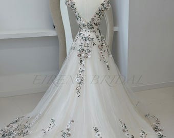 May include: A floor-length off-white bridal gown featuring a deep V-back, cap sleeves, and a full tulle skirt. The dress is adorned with intricate floral embroidery in shades of green, white, and soft pink, with the text Eiren Bridal visible.