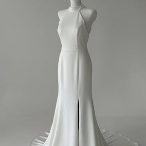 May include: A white bridal gown with a high neck and a front split, displayed on a mannequin. The dress has a fitted bodice and a flowing skirt that extends into a train. The background is plain. The gown is floor-length.